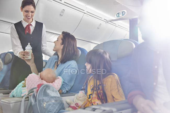 Un agent de bord souriant apporte un biberon à la mère avec bébé dans l'avion — Photo de stock