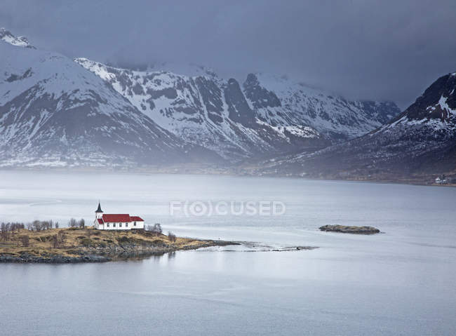 Iglesia remota a lo largo del fiordo frente al mar debajo de las montañas nevadas, Sildpoinesnet, Austvagoya, Noruega - foto de stock
