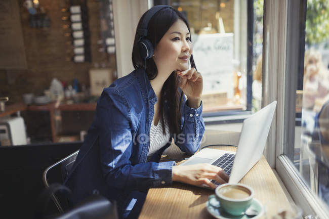 Молода жінка слухає музику з навушниками на ноутбуці і п'є каву у вікні кафе — стокове фото
