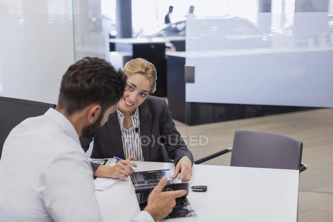 Smiling car saleswoman showing brochure to male customer in car dealership office — Stock Photo