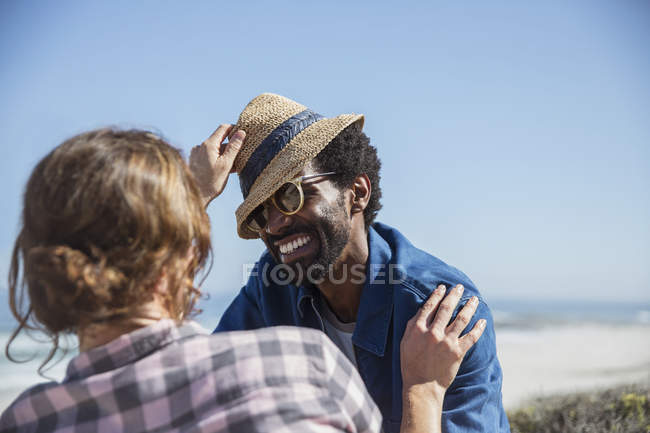 Verspieltes multiethnisches Paar mit Hut am sonnigen Sommerstrand — Stockfoto
