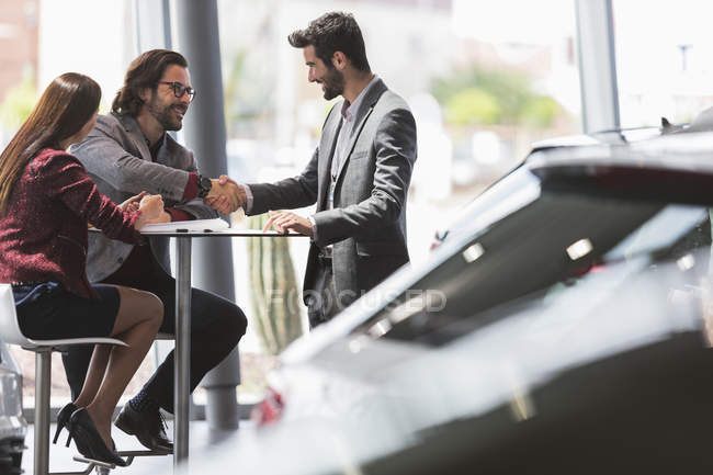 Car salesman handshaking with couple customers at table in car dealership showroom — Stock Photo