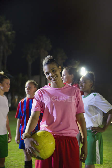 Retrato sorridente, confiante jovem jogadora de futebol feminino com bola em campo à noite — Fotografia de Stock