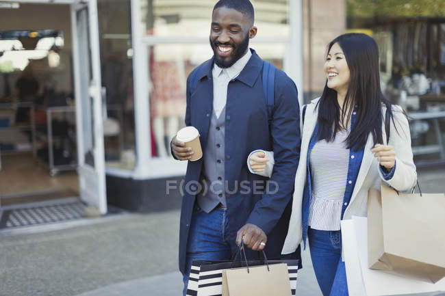 Sorridente giovane coppia che cammina a braccetto lungo il negozio con caffè e shopping bags — Foto stock