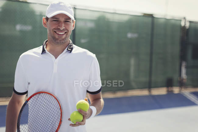 Портрет впевнений, усміхнений молодий тенісист тримає тенісні ракетки і тенісні м'ячі на сонячному тенісному корті — стокове фото