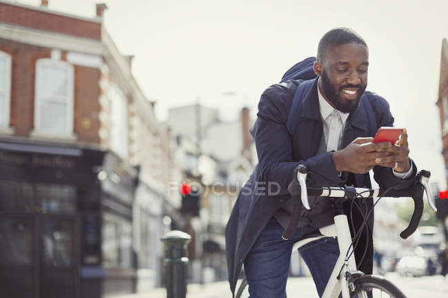 Young businessman commuting with bicycle, texting with cell phone on sunny urban street — Stock Photo