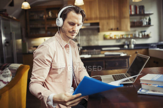 Mann mit Kopfhörern arbeitet am Laptop, liest Papierkram in der Küche — Stockfoto