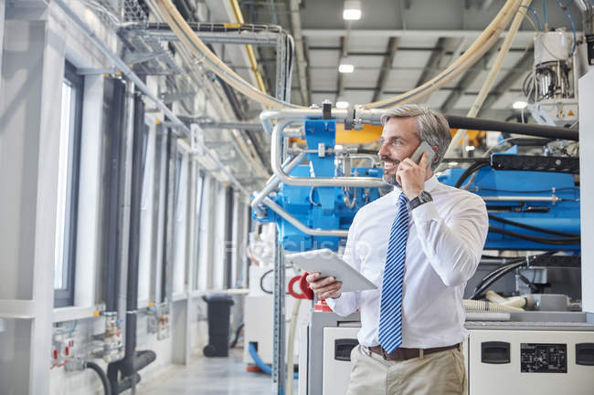 Male supervisor with digital tablet talking on cell phone next to plastic industrial molder machinery in factory — Stock Photo