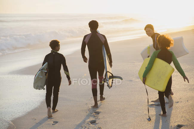 Familia multiétnica que lleva tablas de surf y boogie en la soleada playa del atardecer de verano - foto de stock