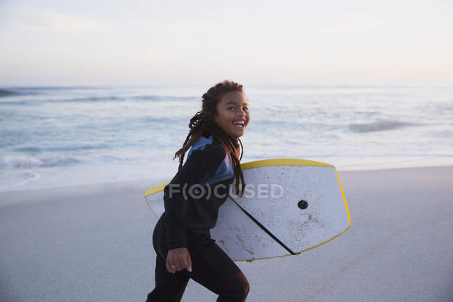 Portrait happy pre-adolescent girl running with boogie board on summer beach at dusk — Stock Photo