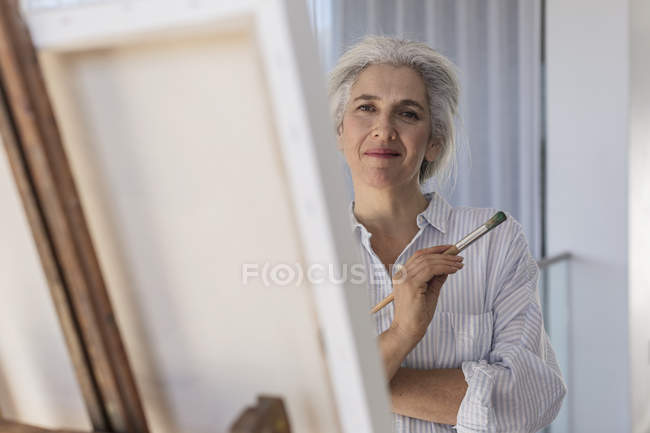 Portrait confident mature woman painting at canvas on easel — Stock Photo