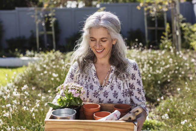 Улыбающаяся взрослая женщина с подносом для садоводства в солнечном саду — стоковое фото
