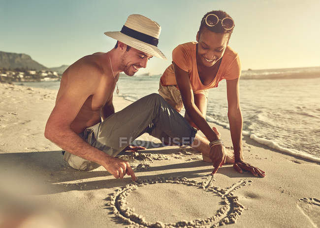 Young couple drawing in sand on sunny summer ocean beach — Stock Photo