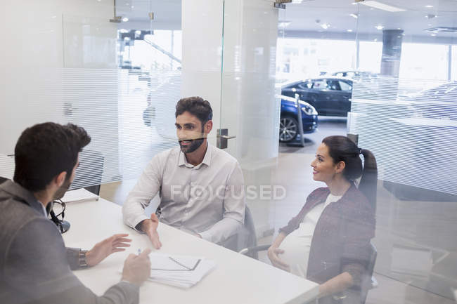 Вагітна пара розмовляє з продавцем автомобілів, обговорюючи фінансові документи в автосалоні — стокове фото