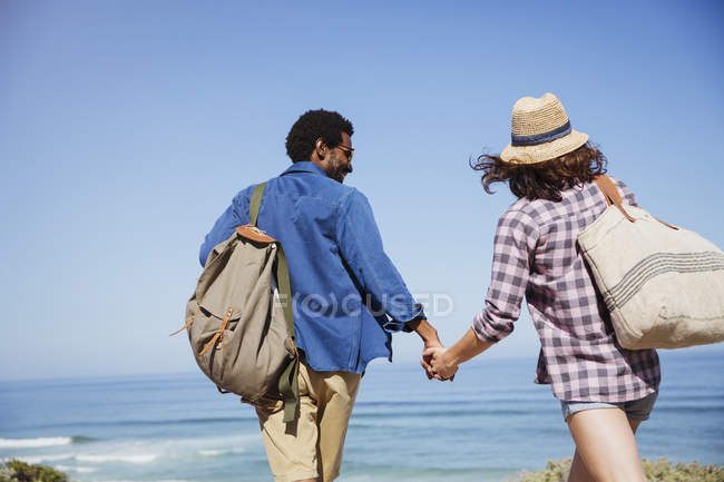Прихильна багатоетнічна пара тримається за руки, гуляючи на сонячному літньому океані — стокове фото