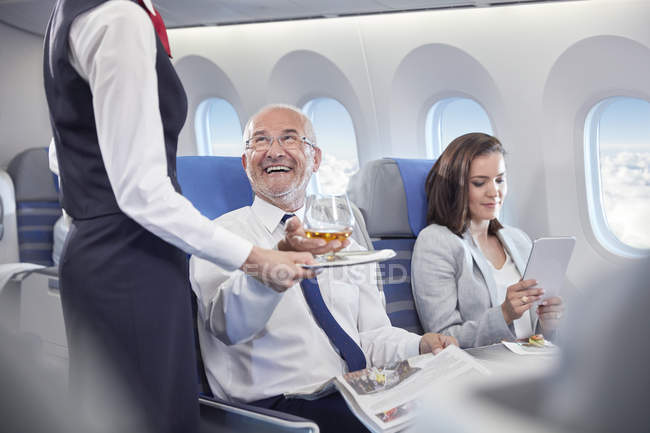 Asistente de vuelo sirviendo whisky a un hombre de negocios sonriente cabalgando en primera clase en un avión - foto de stock