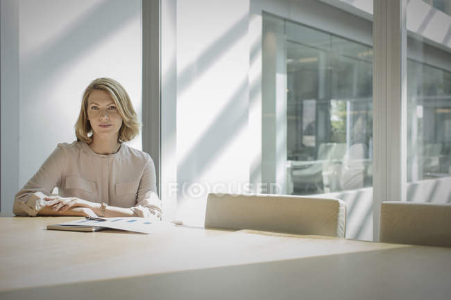 Retrato mujer de negocios confiada sentada en la mesa de conferencia soleada - foto de stock