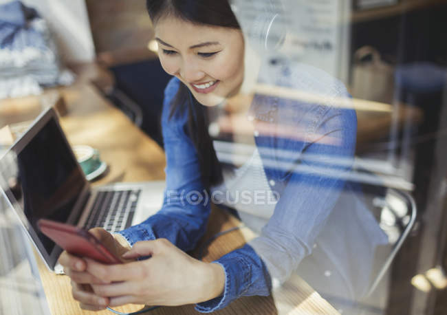 Mujer joven sonriente escuchando música con auriculares y mensajes de texto con teléfono celular en la computadora portátil en la ventana de la cafetería - foto de stock