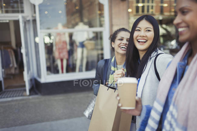 Mulheres risos amigos andando ao longo da frente de loja com café e sacos de compras — Fotografia de Stock