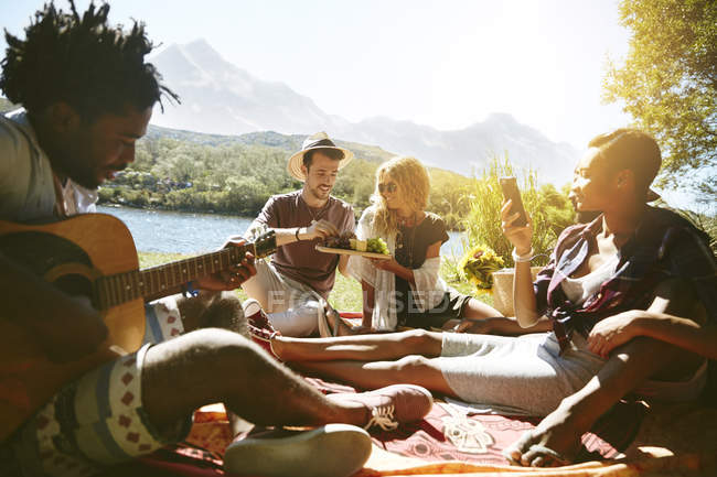 Молодые друзья тусуются, играют на гитаре и наслаждаются пикником у солнечного летнего берега — стоковое фото