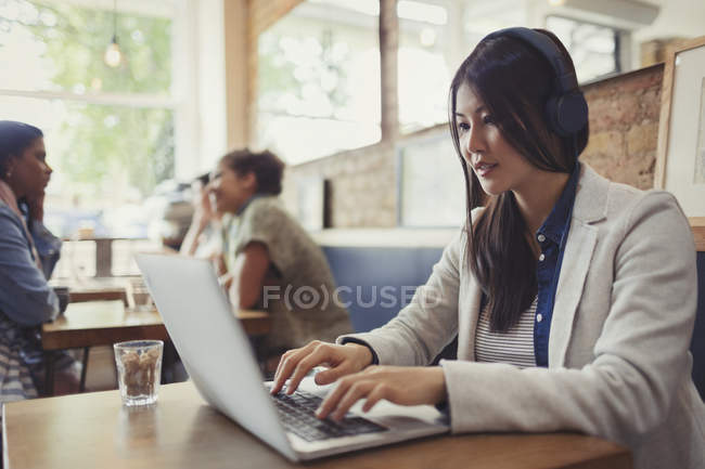Young woman with headphones using laptop at cafe table — Stock Photo