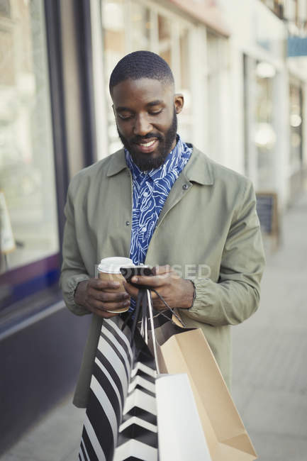 Jeune homme souriant avec café et sacs à provisions textos avec téléphone portable sur le trottoir urbain — Photo de stock