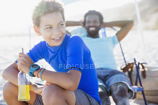 Sorridente padre e figlio bere succo di frutta, rilassante sulla spiaggia estiva soleggiata — Foto stock