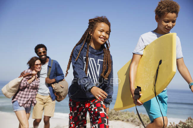 Família sorridente carregando prancha de boogie, andando no caminho ensolarado da praia de verão — Fotografia de Stock