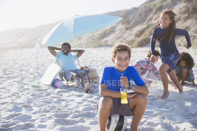 Ritratto sorridente ragazzo pre-adolescente bere succo di frutta sulla spiaggia estiva soleggiata con la famiglia — Foto stock