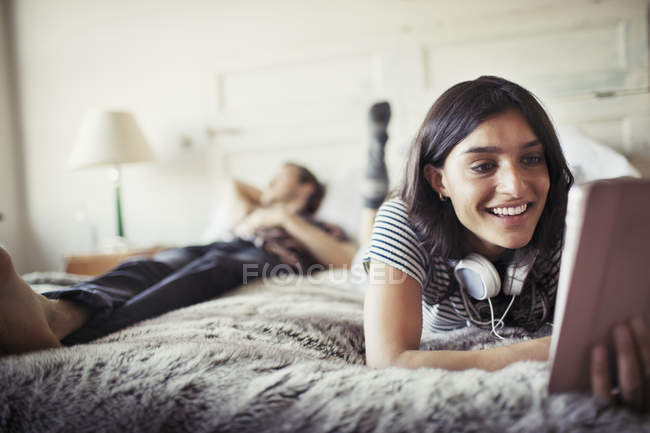 Giovane donna sorridente con le cuffie utilizzando tablet digitale sul letto — Foto stock