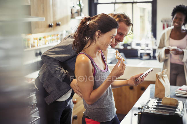 Sorrindo casal mensagens de texto com telefone inteligente, comer torrada na cozinha — Fotografia de Stock