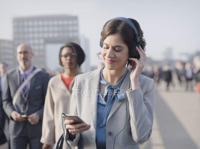 Femme d'affaires souriant, écoutant de la musique avec écouteurs et téléphone intelligent sur la passerelle piétonne urbaine — Photo de stock
