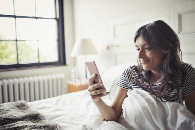 Молода жінка розслабляється в ліжку, смс зі смартфоном — стокове фото