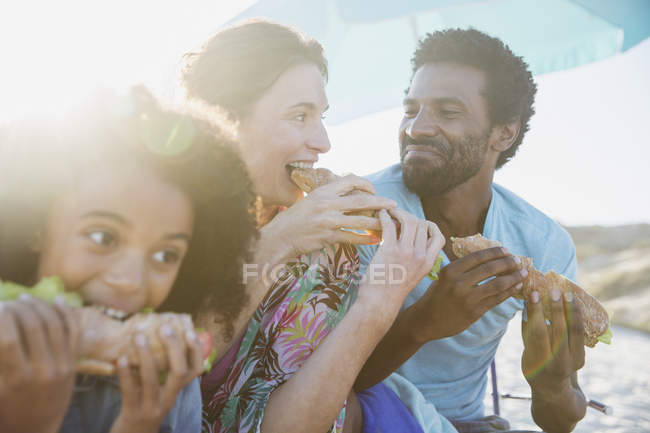 Happy family eating baguette sandwiches on sunny summer beach — Stock Photo