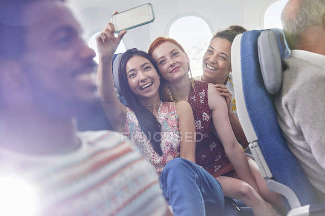 Young women friends with camera phone taking selfie on airplane — Stock Photo
