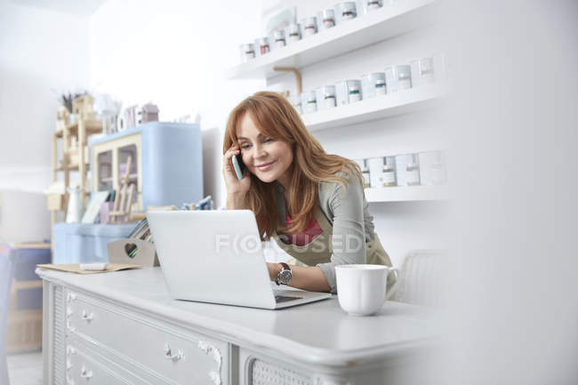 Propietaria sonriente del negocio femenino usando el ordenador portátil y hablando por teléfono celular en el mostrador en la tienda de pintura de arte - foto de stock