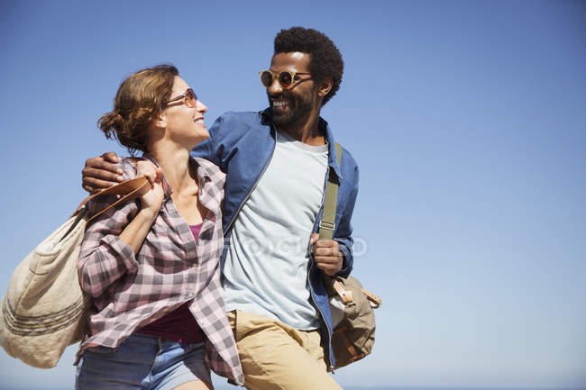 Affectionate multi-ethnic couple hugging and walking below sunny summer blue sky — Stock Photo