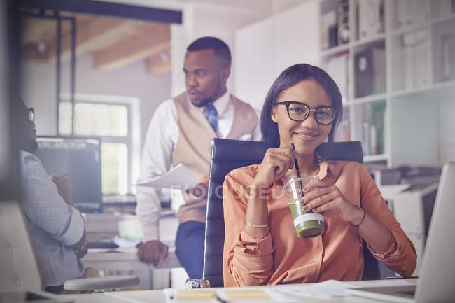 Портрет улыбающейся уверенной бизнесвумен пьет зеленый смузи в офисе — стоковое фото
