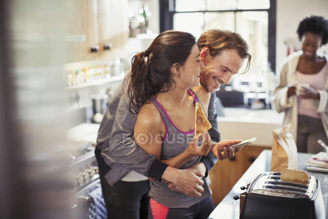 Coppia giocosa che si abbraccia, messaggia con lo smartphone e mangia pane tostato — Foto stock