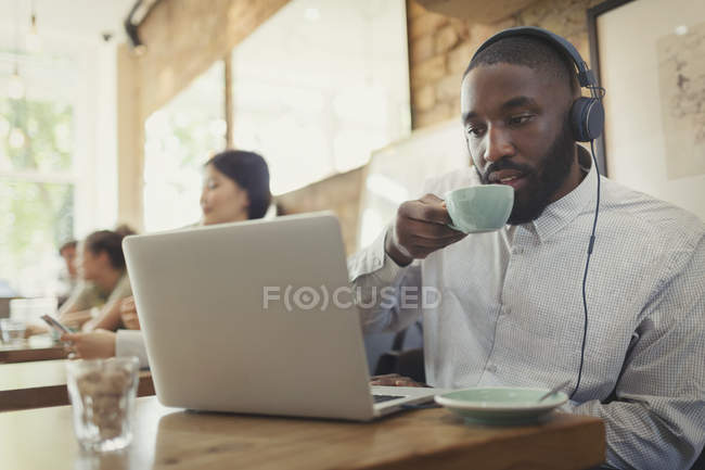 Homme avec écouteurs utilisant un ordinateur portable et boire du café dans le café — Photo de stock