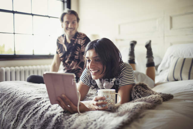 Улыбающаяся пара расслабляется, пьет кофе и использует цифровой планшет на кровати — стоковое фото
