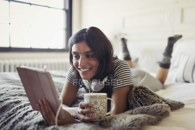 Усміхнена молода жінка п'є каву і використовує цифровий планшет на ліжку — стокове фото