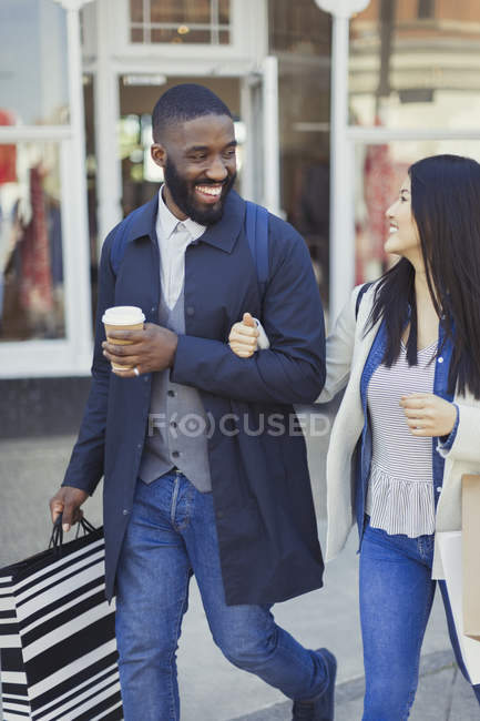 Sonriente pareja joven caminando por el escaparate con café y bolsas de compras - foto de stock