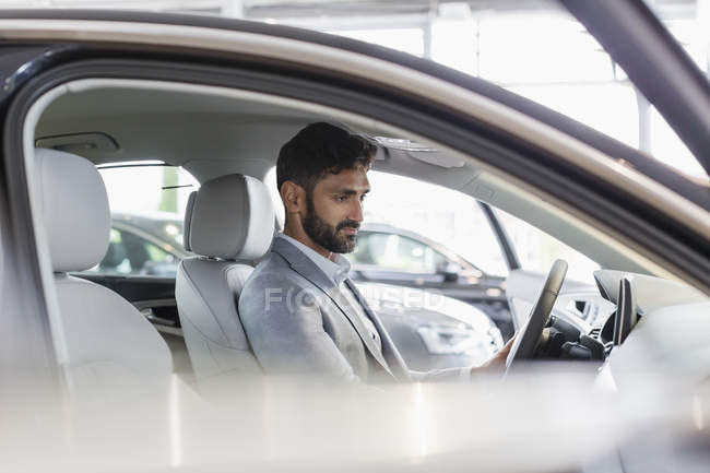 Cliente masculino sentado en el asiento del conductor del coche nuevo en la sala de exposición de concesionarios de automóviles - foto de stock