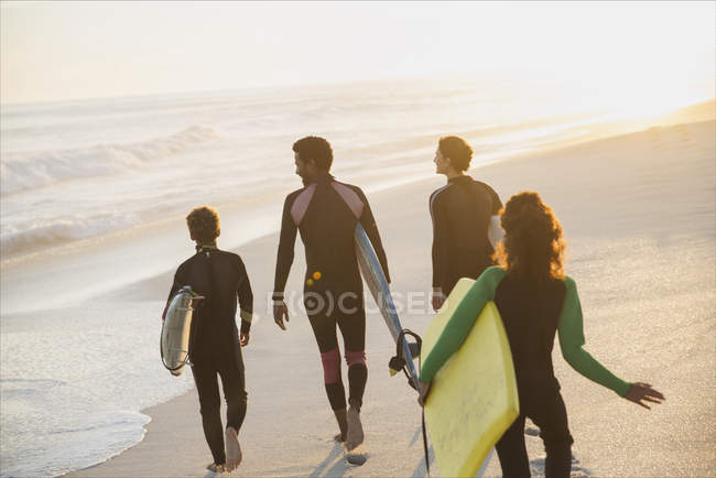 Familia caminando, llevando tablas de surf y boogie board en la soleada playa del atardecer de verano - foto de stock