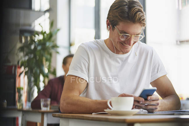 Усміхнений чоловік з навушниками за допомогою мобільного телефону і пиття кави в кафе — стокове фото
