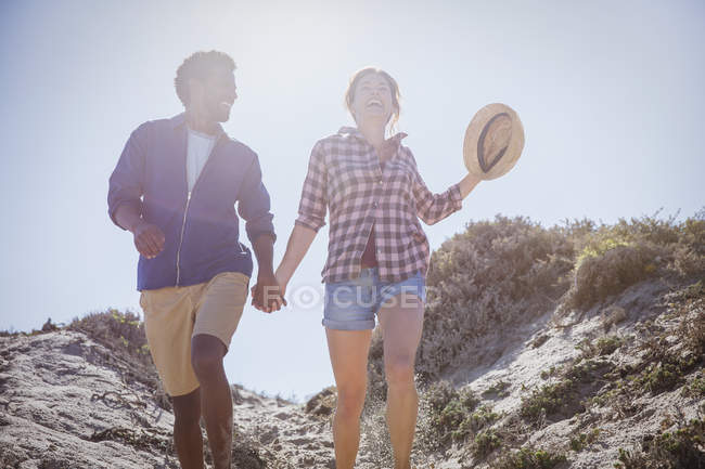 Pareja multiétnica caminando, tomados de la mano en el soleado camino de playa de verano - foto de stock