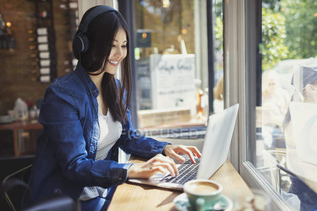 Young woman with headphones using laptop at sunny cafe window — Stock Photo