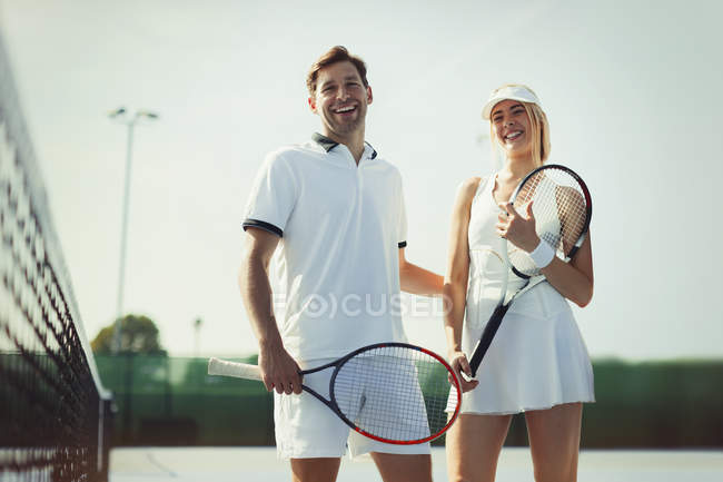 Portrait de joueurs de tennis souriants et confiants tenant des raquettes de tennis sur un court de tennis ensoleillé — Photo de stock