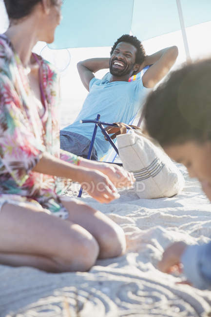 Улыбающийся мужчина расслабляется с руками за головой на летнем пляже с семьей — стоковое фото